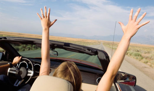 A young couple traveling in a red convertible. The passenger raises her two arms as they travel down a county highway.