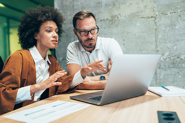 Two coworkers having a conversation while looking and pointing at a laptop.