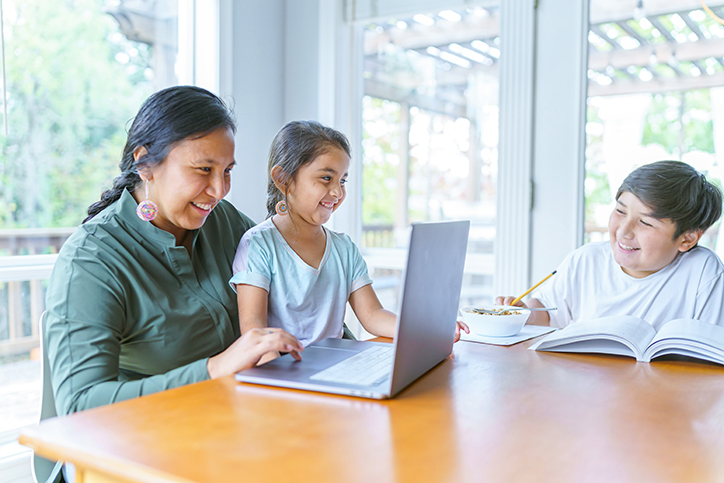 Mother and two children spending time at a dinner table while she works on a laptop.