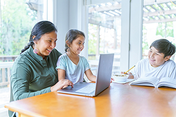 Mother and two children spending time at a dinner table while she works on a laptop.