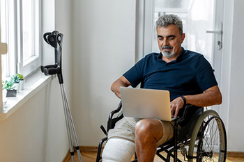 Man sitting in a wheelchair with a leg cast, looking at his laptop.