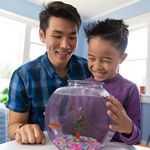 A young boy and his dad are at home and smiling at a fish in a fish bowl. 