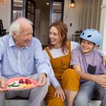 An older man sits on the steps of his front porch next to his daughter and grandson. 